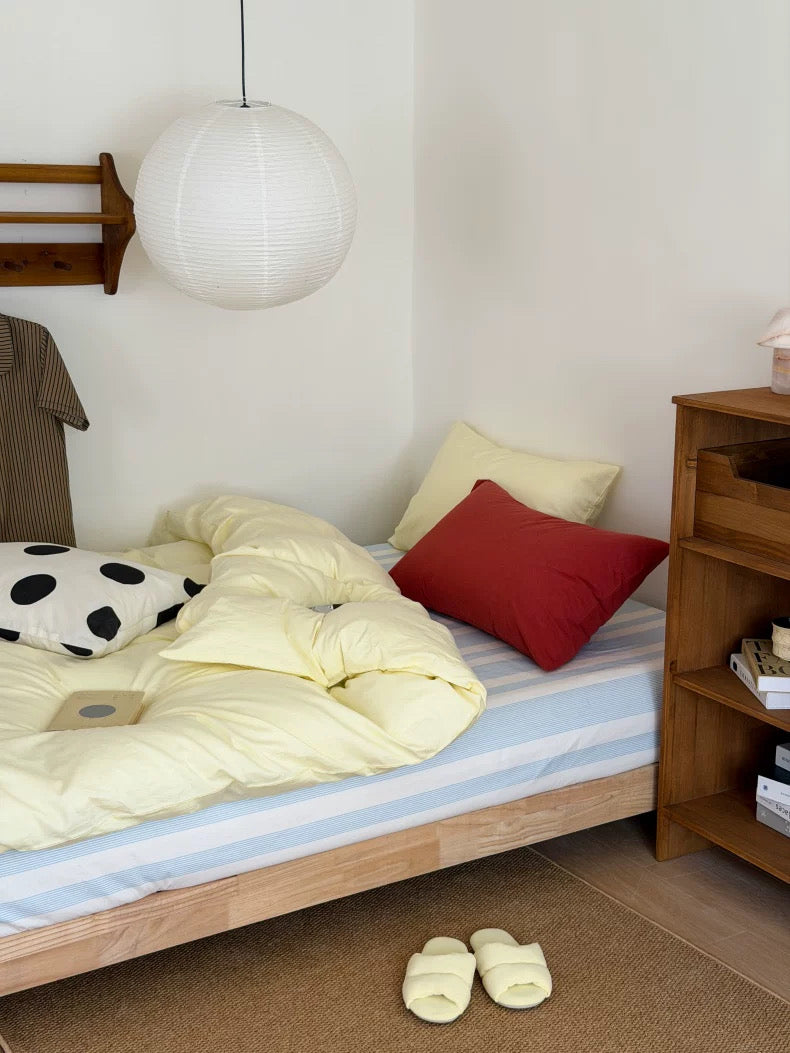 A cozy bedroom corner with a light wood bed frame, pale yellow bedding, blue and white striped sheets, a red accent pillow, a black-and-white polka dot pillow, a white hanging pendant lamp, and a wooden nightstand.