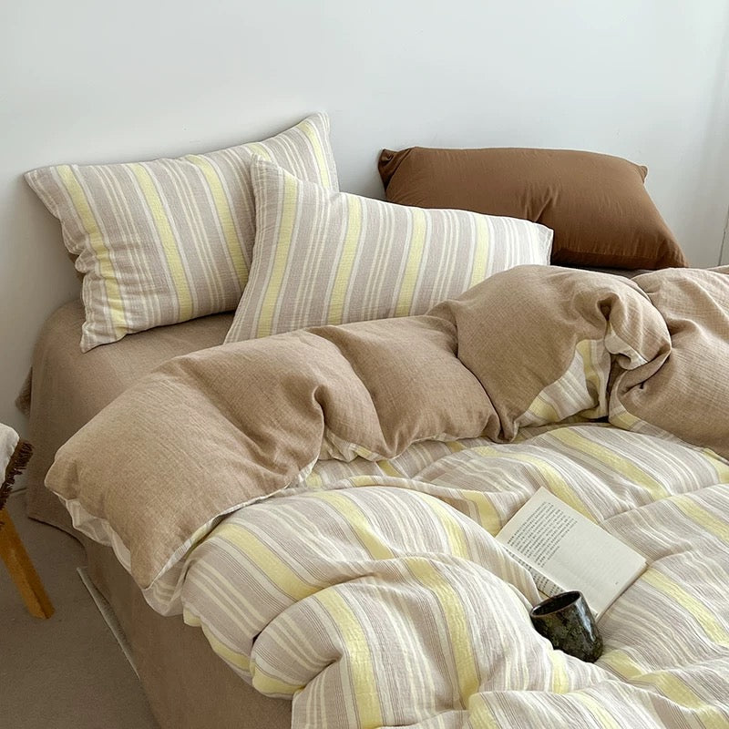A cozy bed styled with a beige and white striped bedding set, including two striped pillows, a matching duvet cover, and solid beige pillows, set against a plain white wall.