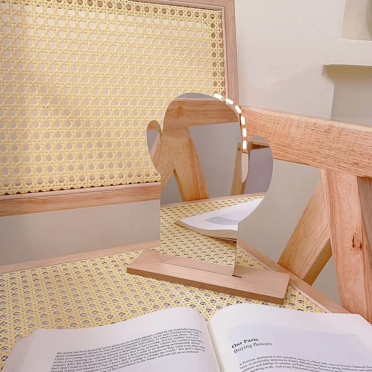 A small wooden tabletop mirror with a stand, shaped like a stylized figure, placed on a woven yellow and white tablecloth next to an open book. A larger cane-rattan screen and wooden chair are visible in the softly lit room.
