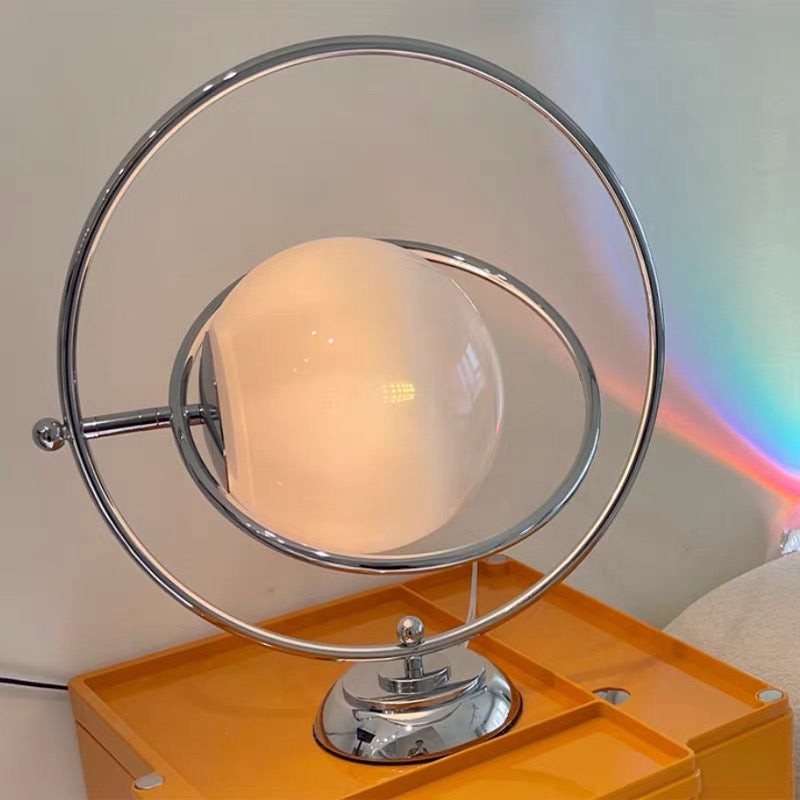 A modern table lamp with a spherical frosted white glass shade and a chrome metal frame, resting on an orange side table against a light wall.
