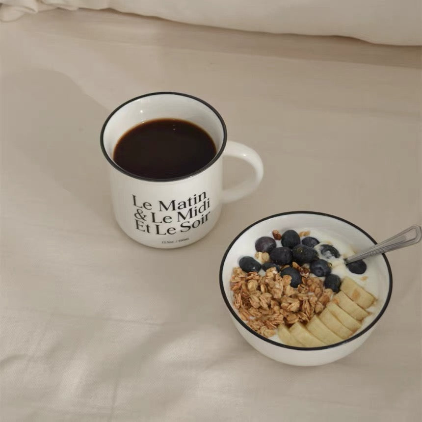 A white ceramic mug with black text reading 'Le Matin & Le Midi Et Le Soir' sits on a beige fabric surface next to a white bowl of breakfast cereal with blueberries, granola, and banana slices.