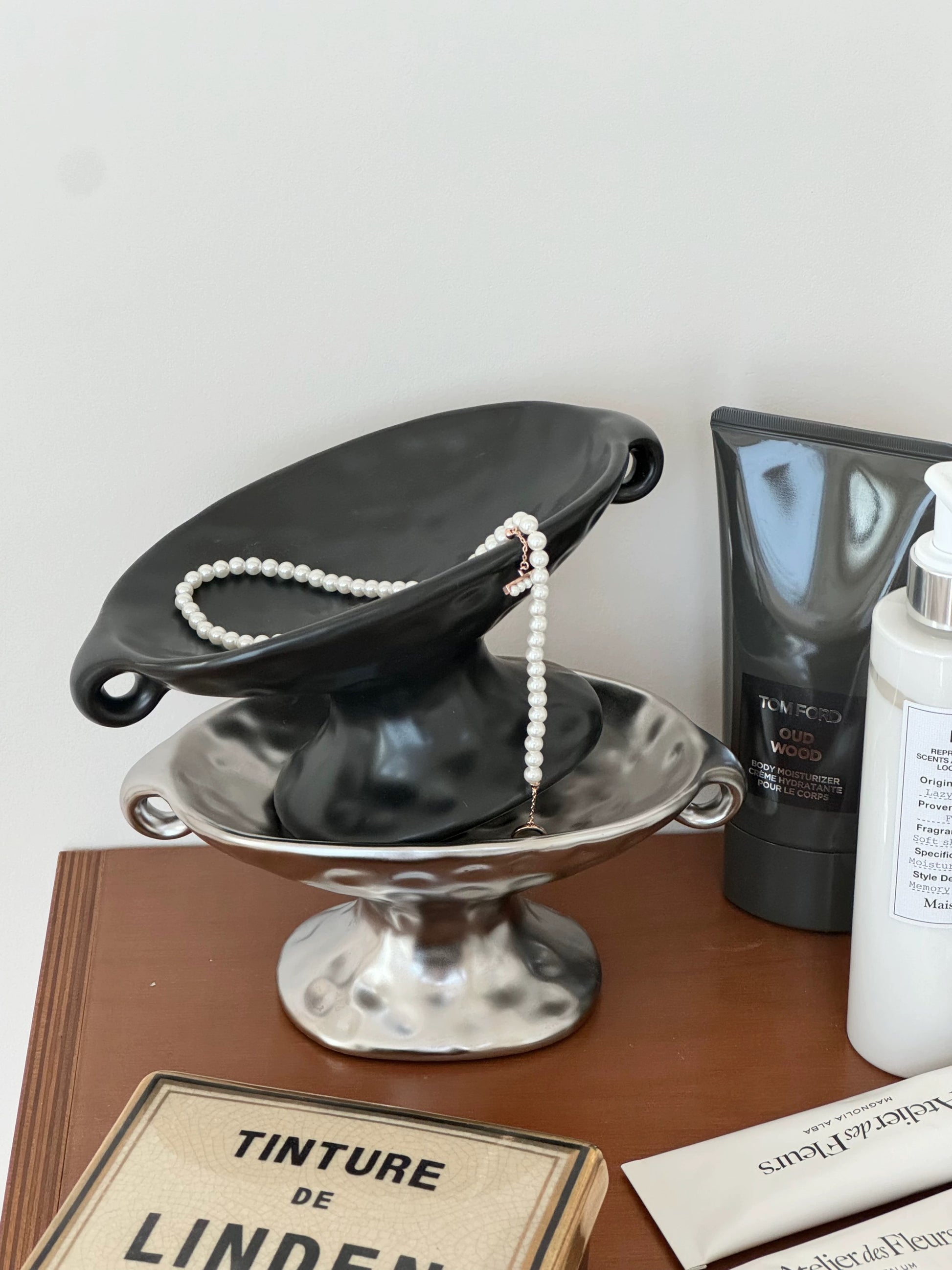 A black ceramic jewelry display stand shaped like a bowl, holding a strand of white pearls, styled on a wooden surface next to skincare products including a black tube and a white bottle.