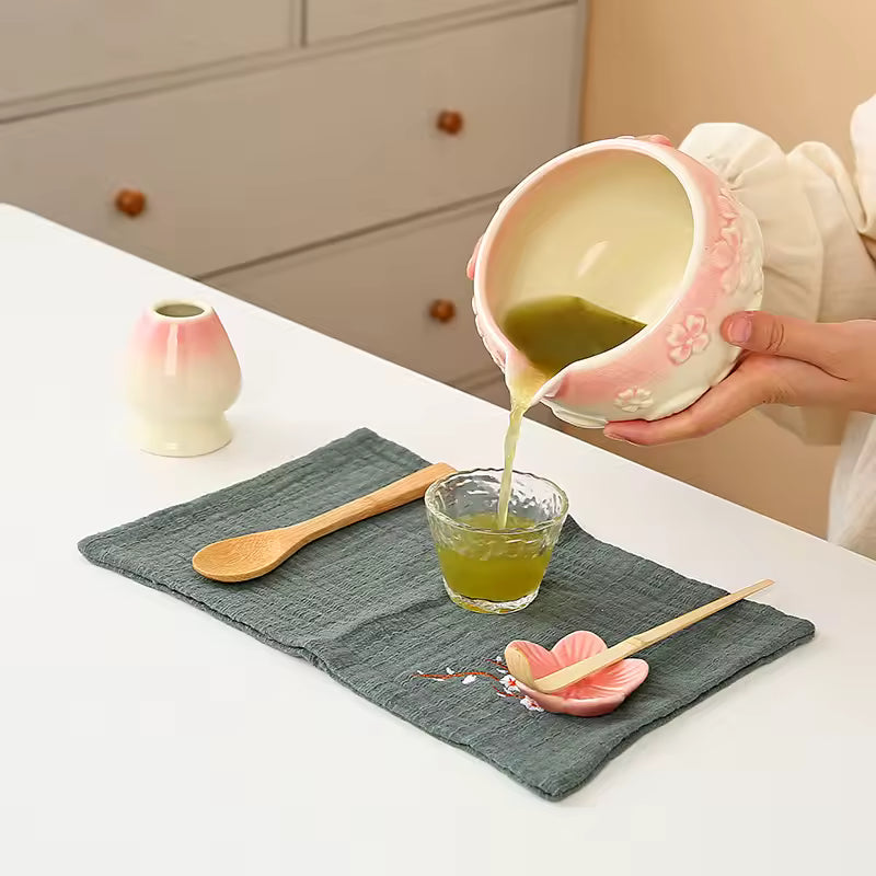 Person pouring green liquid from a ceramic pitcher into a small glass bowl on a white table.