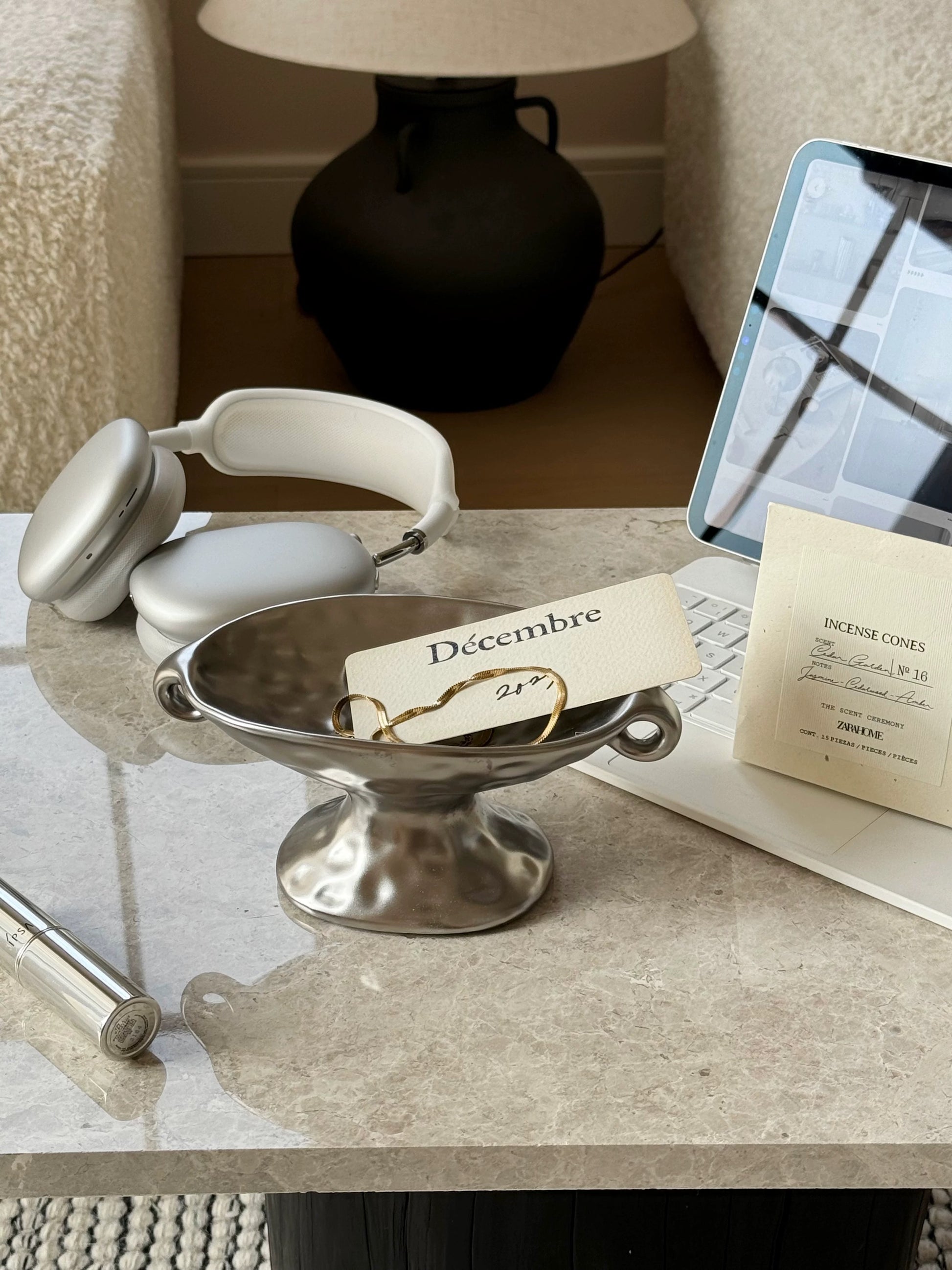A styled tabletop scene showing a pair of silver over-ear headphones, a silver pedestal bowl with a small card reading “Décembre” and a gold ring, a laptop, and a cream card on a marble-like surface in an indoor room.