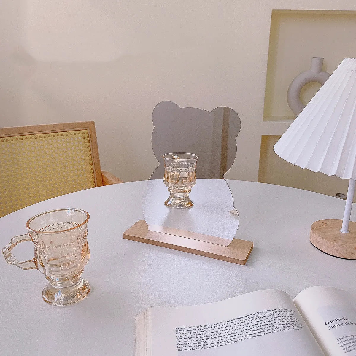 A small clear glass candle holder with a flared base sits on a wooden base on a white table, styled in a warm, minimalist room with a white pleated table lamp, a translucent pink glass mug, and an open book.