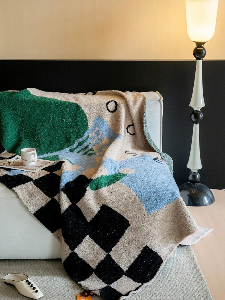 A textured knit throw blanket with a geometric pattern in beige, black, green, and light blue, draped over a white sofa in a modern living room setting next to a lit floor lamp.