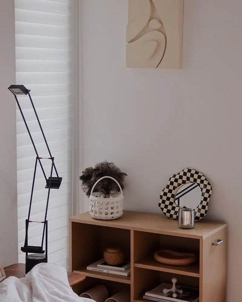 A styled bedroom corner featuring a light wood cubby shelf with decorative items, a black-and-white checkered mirror, a white basket with dried grass, and a modern black floor lamp next to white window blinds.