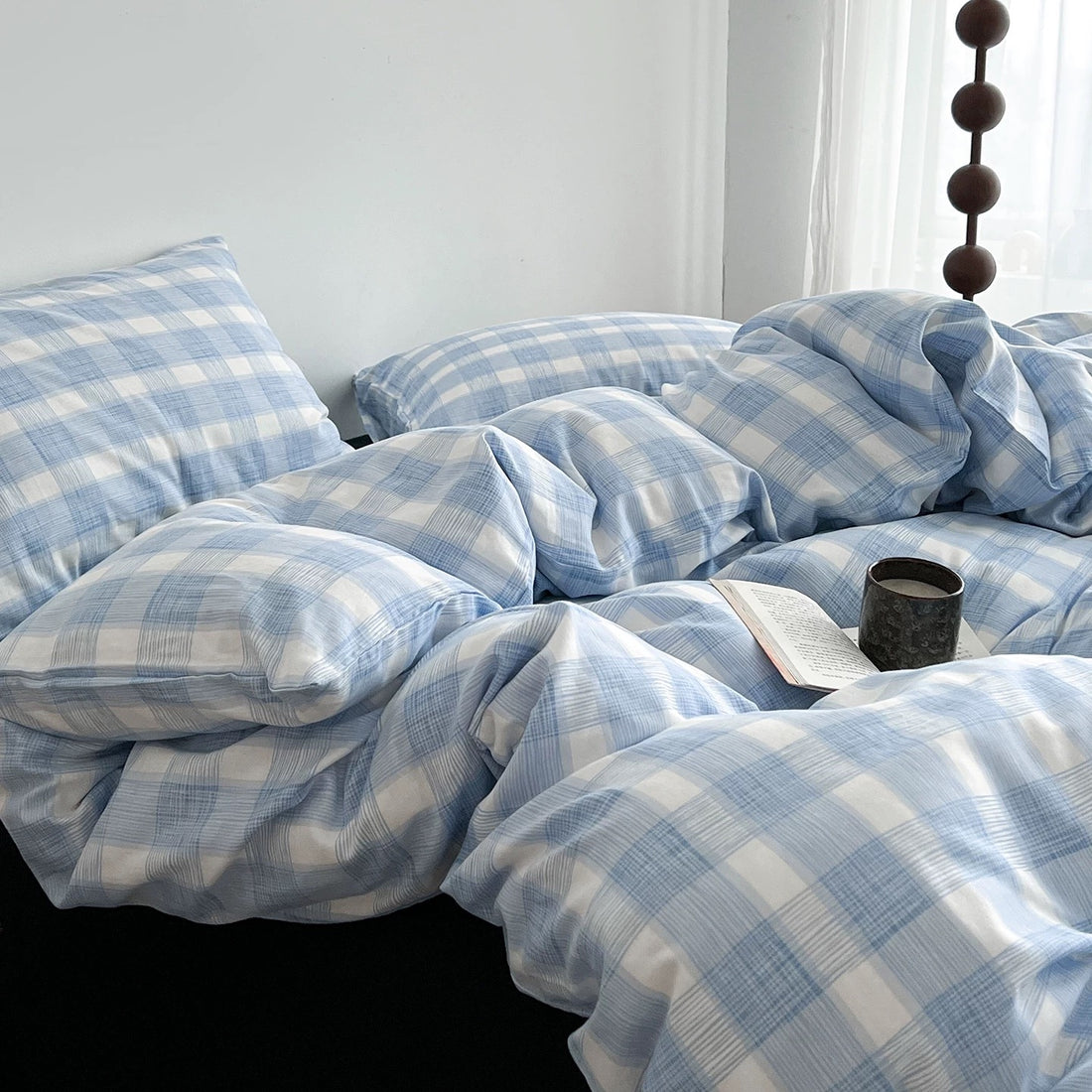 Light blue and white plaid bedding set with matching pillowcases and a duvet cover, styled on a bed with dark pillows and a book resting on the duvet.