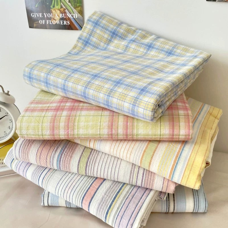 A stack of four folded plaid and striped fabric pieces in soft pastel colors, including blue, pink, yellow, and white, arranged on a light surface indoors.