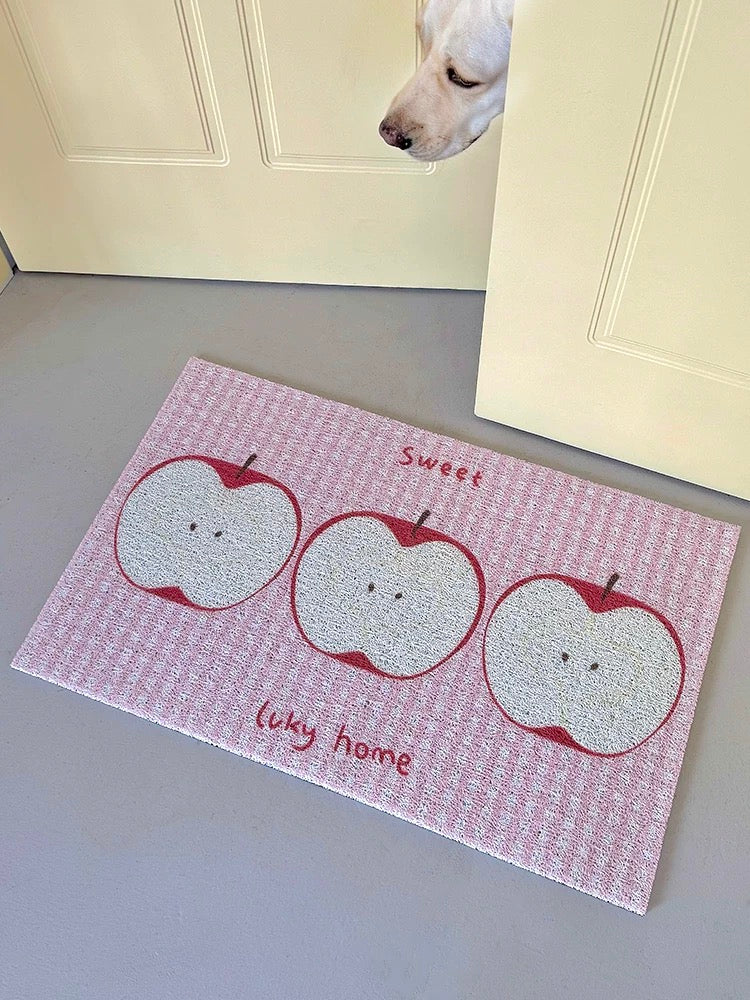 A pink and white checkered doormat with three cartoon apple illustrations and the words 'Sweet Lucy home', shown on a gray floor in front of cream-colored doors, with a dog's head peeking from the top right.