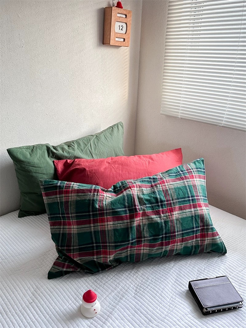 A Christmas pillowcase in green and red tartan sits beside a matching plaid design, with solid red and green pillowcases behind them.