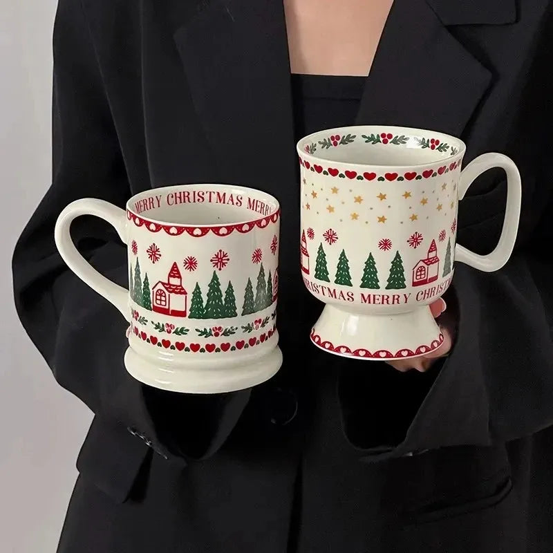 Two white ceramic Christmas-themed mugs with red and green holiday patterns, held by a person wearing a black long-sleeve top against a light gray background.