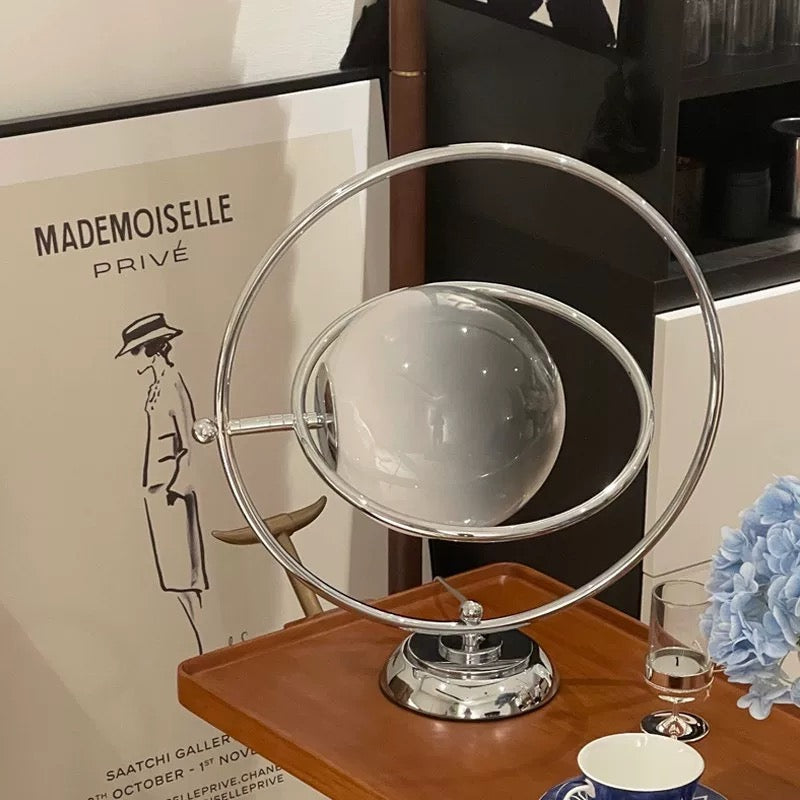 A modern round vanity mirror with a chrome metal frame and a glossy white spherical glass cover, displayed on a wooden tray on a tabletop with decorative props.
