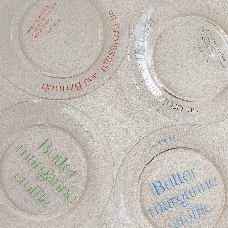Three clear glass plates with French text are arranged on a textured cream surface. One plate has red text, one has blue text, and one has green text.