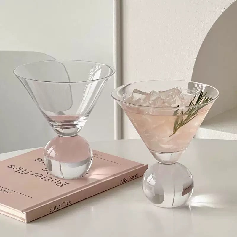 Clear glass with pink cocktail and ice cubes on a white surface with a book underneath.