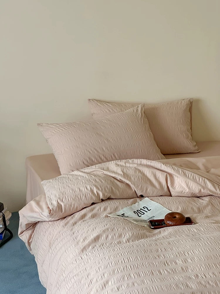 A bed dressed in a textured blush pink bedding set with matching pillows, set against a plain beige wall with a small blue floor area visible on the left.