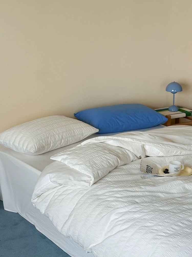 A neatly made bed with white textured bedding, two pillows (one white, one blue), and a small blue table lamp on a side table against a beige wall.