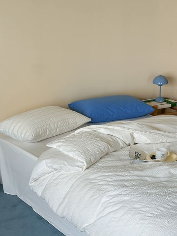 A neatly made bed with white textured bedding, two pillows (one white, one blue), and a small blue table lamp on a side table against a beige wall.