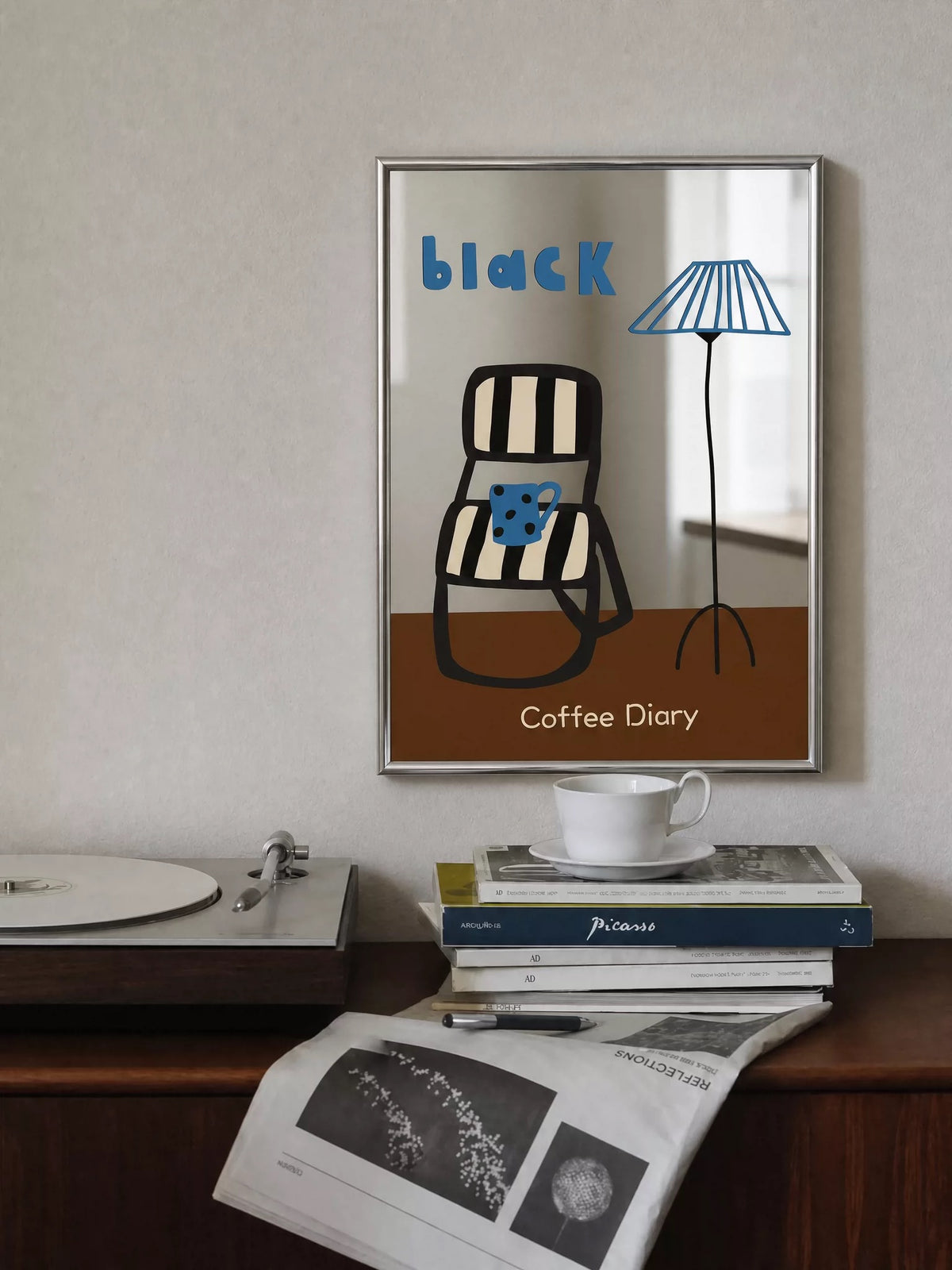 The Coffee Diary wall mirror hangs above a desk, showing a black chair and blue lamp illustration over a brown panel.