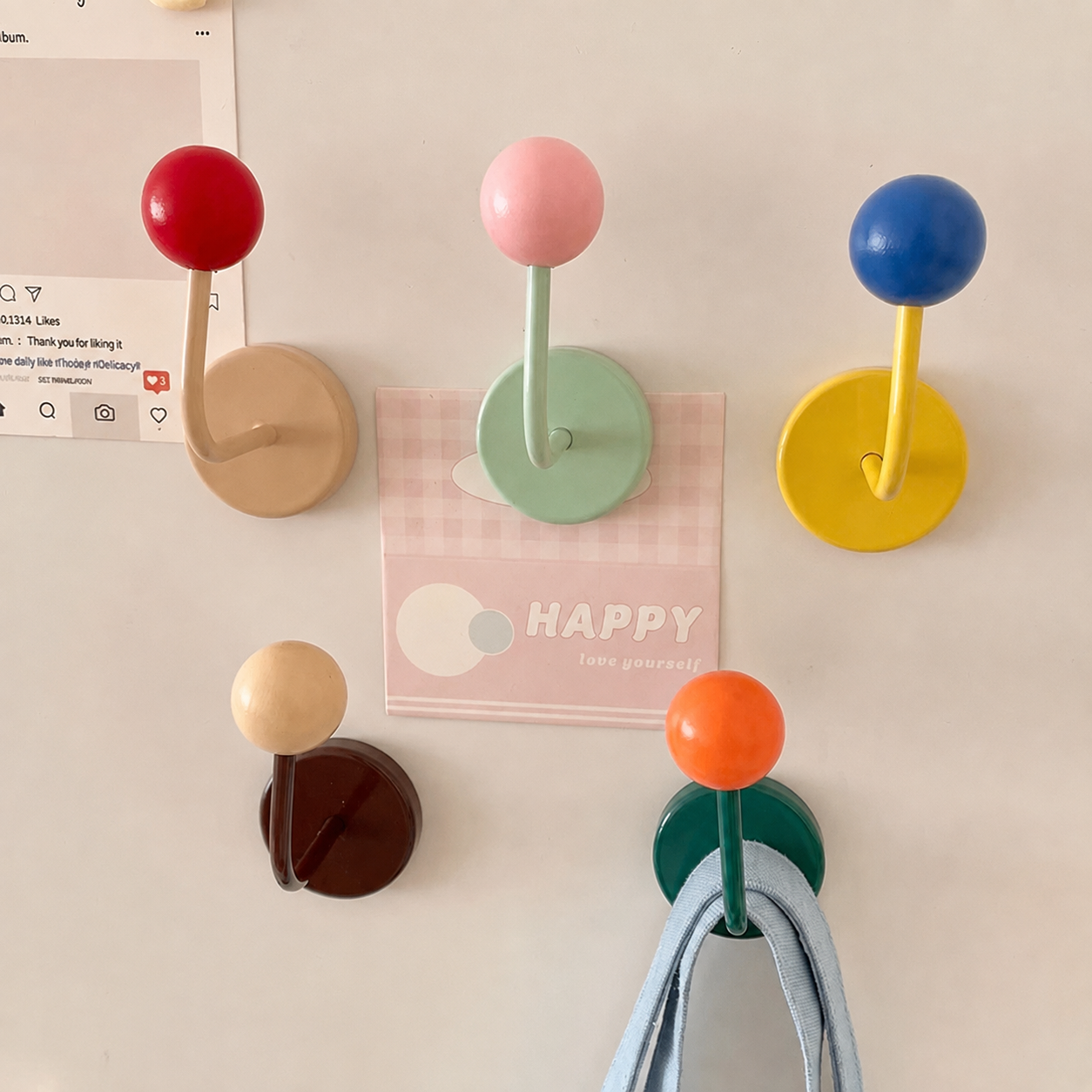 Five colorful wall-mounted coat hooks with round bases and spherical tops in red, pink, blue, beige, and dark brown, arranged on a light wall with a small card and hanging cloth.
