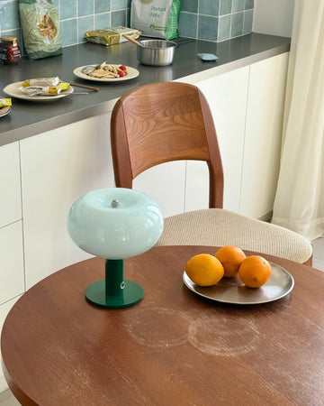 Round wooden table with a decorative lamp and plate of oranges in a kitchen setting.