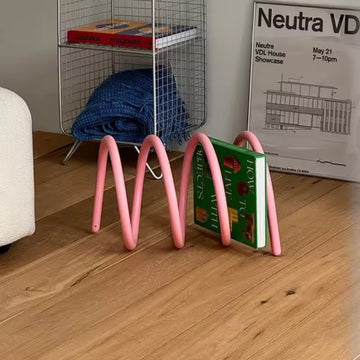 Pink metal bookend holding a green book on a wooden floor with a wire basket in the background.