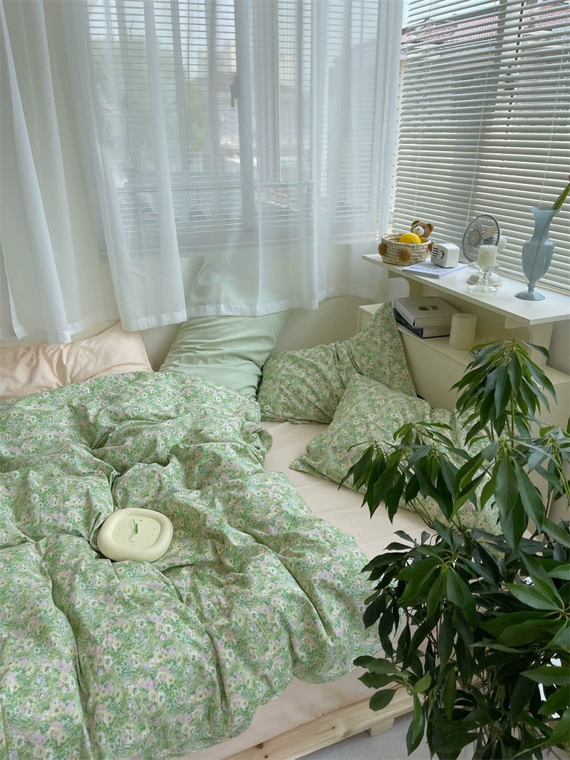 Bedroom with green floral bedding, white curtains, and a small table with items.