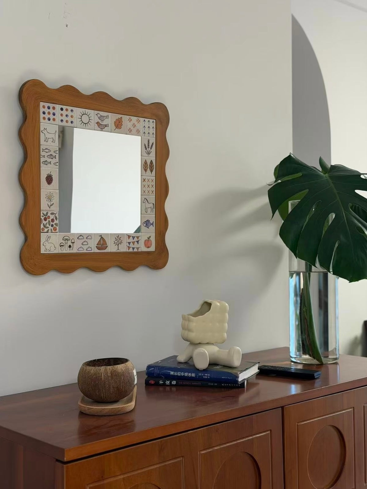 A Cottagecore wall mirror with a wavy brown frame hangs over a cabinet beside a vase of large green leaves.