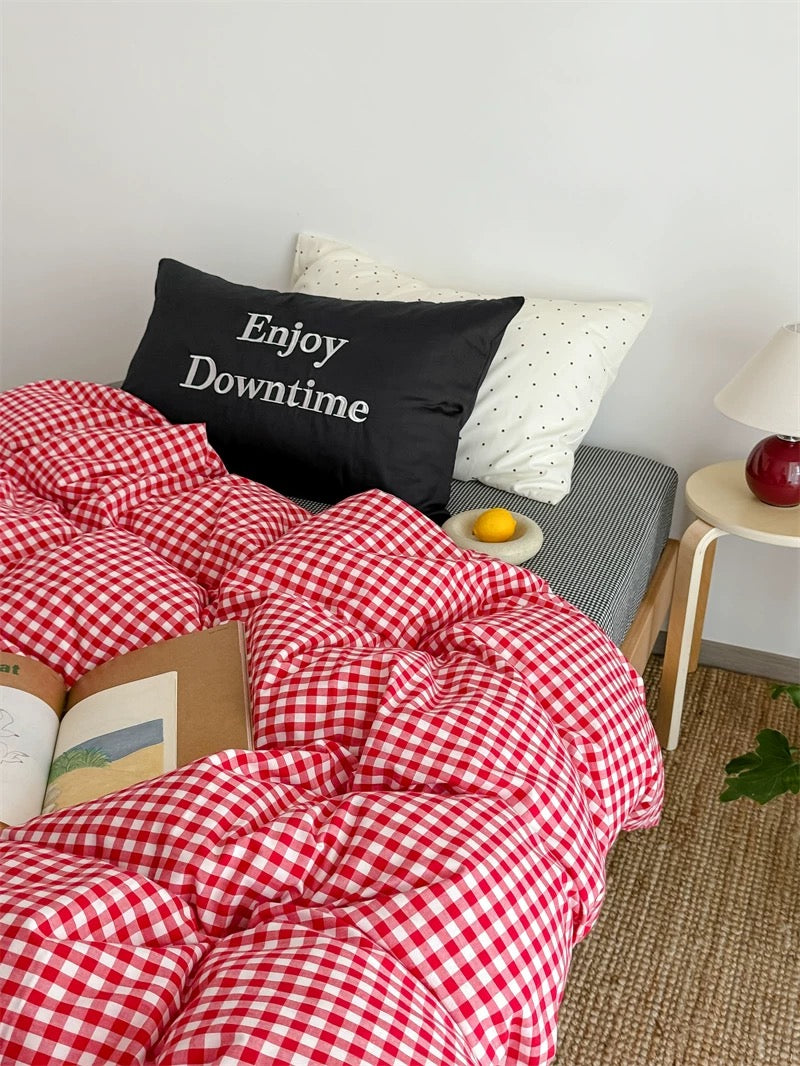 Bed with red checkered comforter, black pillow with 'Enjoy Downtime' text, and small table with fruit.
