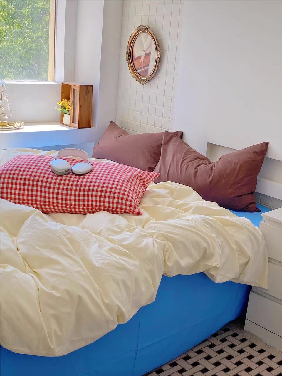 A cozy bed setup in a bright bedroom, featuring a cream-colored duvet, a blue bed frame, a red-and-white gingham pillow, and two dusty rose pillows. A small pair of blue headphones rests on the gingham pillow.