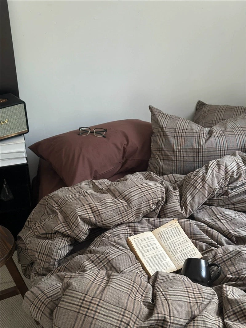 Bed with plaid comforter, pillows, and a book on a wooden floor.