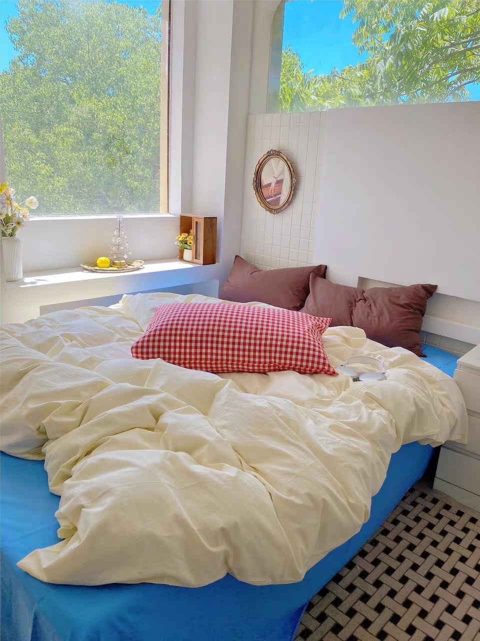 A bed dressed in cream bedding with a red-and-white gingham accent pillow and mauve pillows, set in a bright bedroom with large windows showing green trees outside.