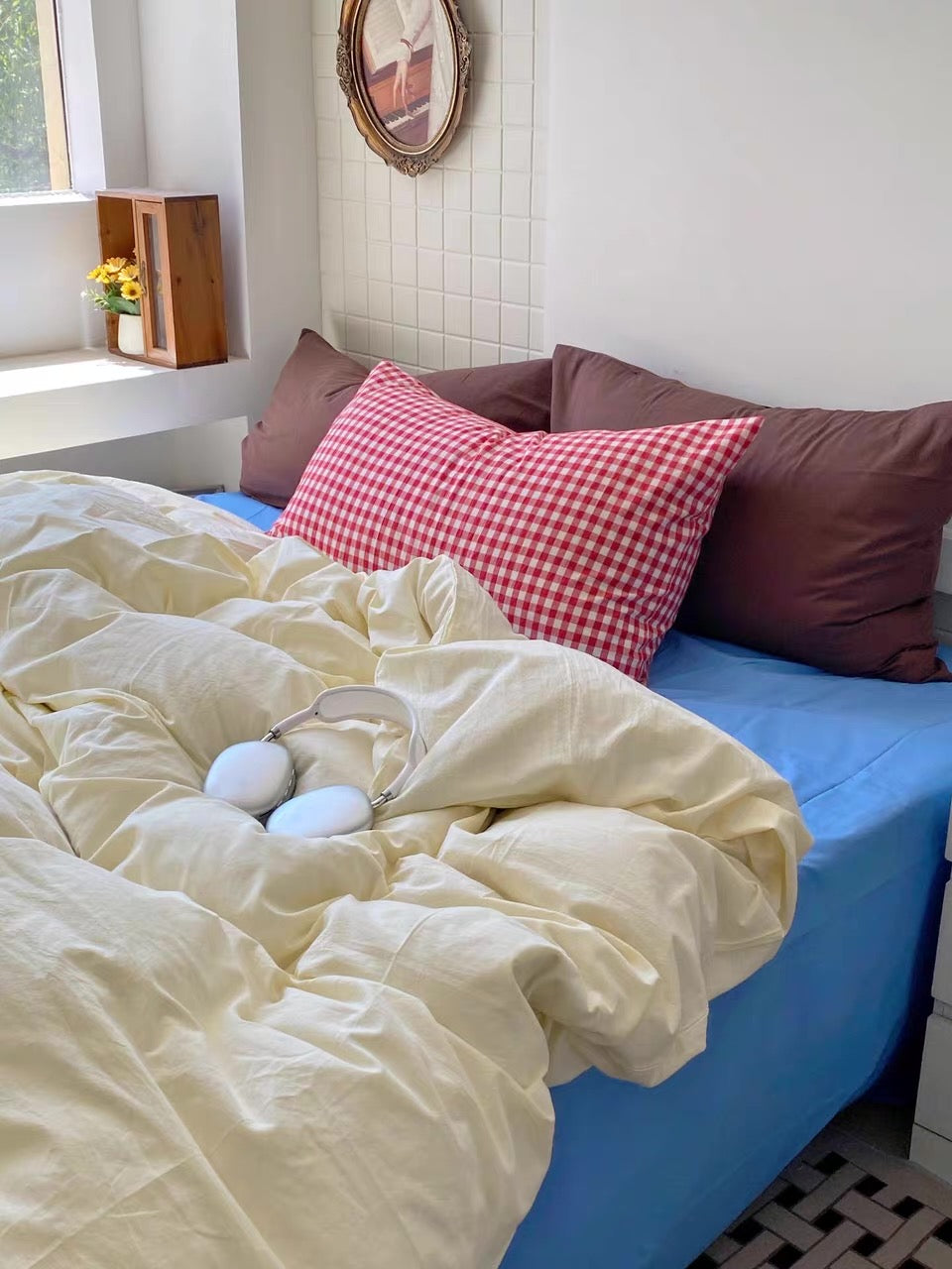 A cozy bedroom scene with a bed made up in soft cream bedding, a bright blue sheet, and several pillows including a red-and-white gingham accent pillow. A pair of white over-ear headphones rests on the duvet.