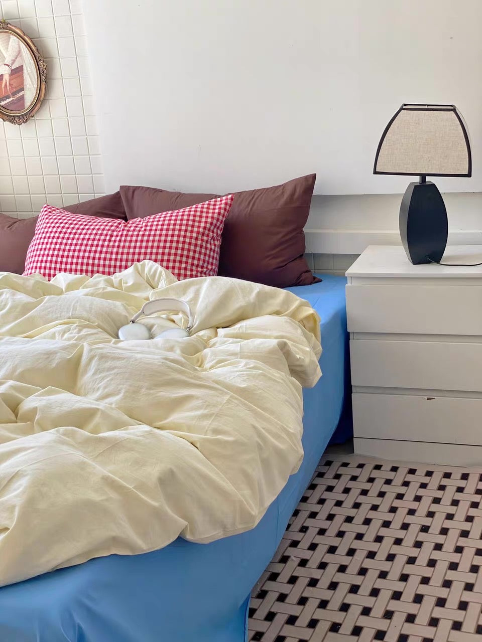 A bed with rumpled cream bedding and a red-and-white gingham accent pillow, styled in a bedroom with a blue bed skirt, white nightstand, black table lamp, and woven rug.