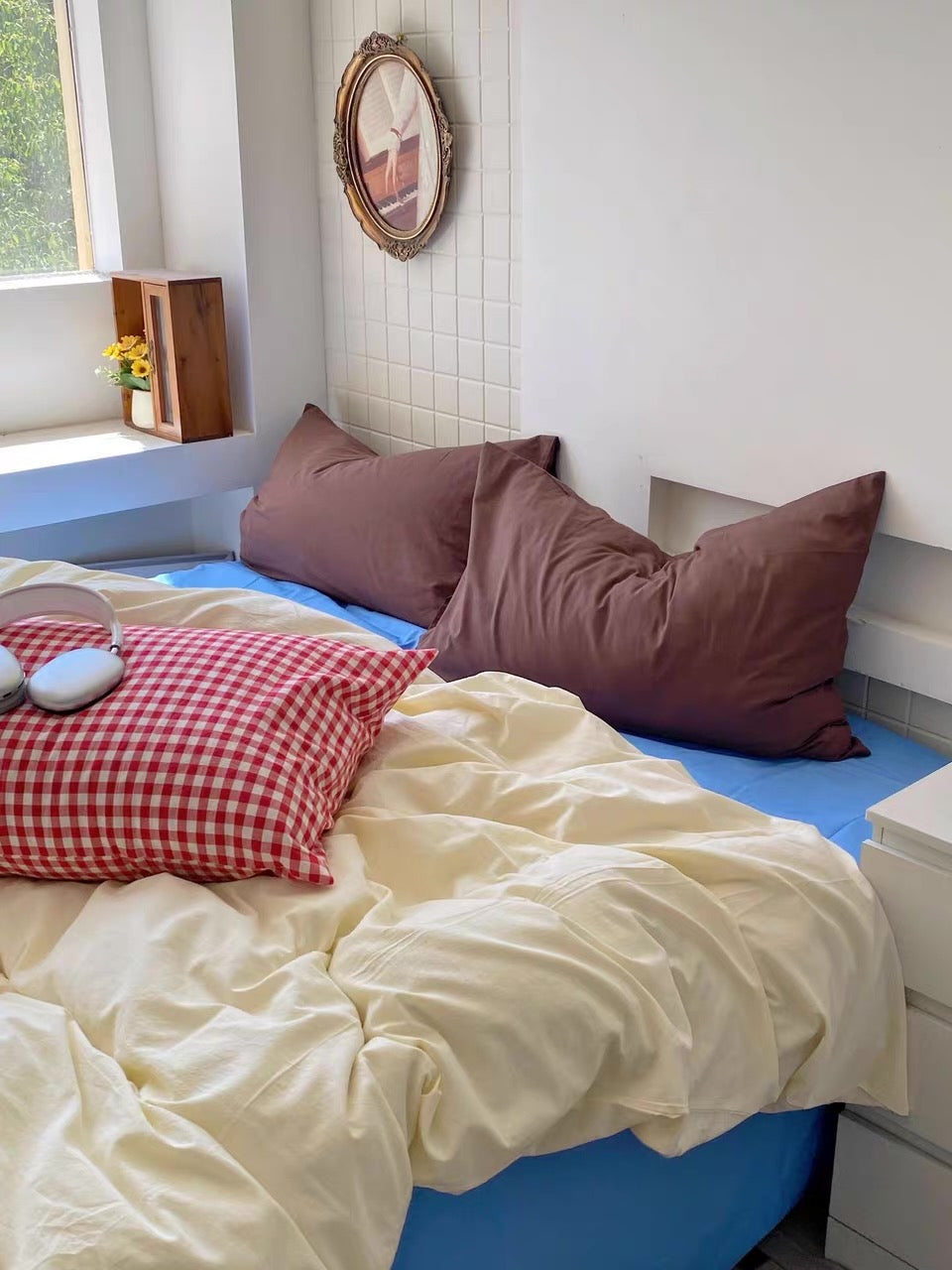 A cozy bed setup in a bright room, featuring cream-colored bedding, a red-and-white gingham accent pillow, and two dusty mauve pillows. A blue mattress or bed frame is visible underneath, with a window, white wall, and small wooden shelf in the background.