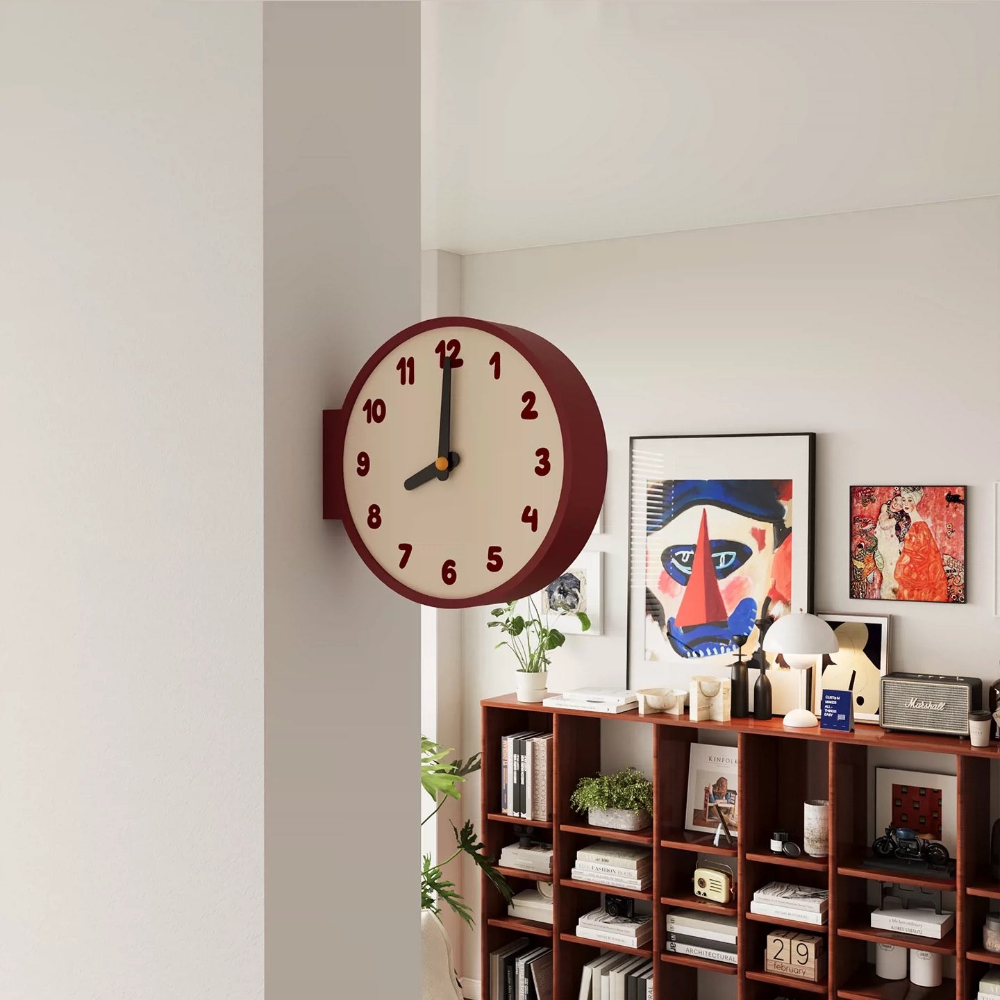 A round wall clock with a cream face, dark red outer rim, and dark red numerals, mounted on a white wall in a modern room with a wooden bookshelf and framed art below.