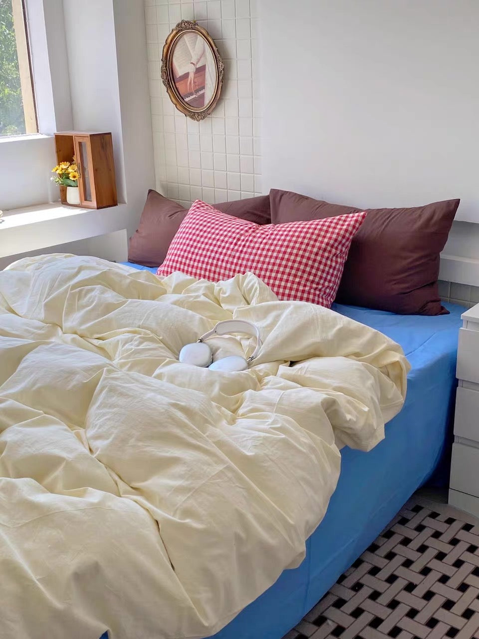 A cozy bedroom scene with a bed made up in a cream-colored duvet cover and matching pillowcases, styled with brown and red gingham accent pillows. The bed is fitted with a bright blue sheet, set against a white tiled wall with a small round mirror.