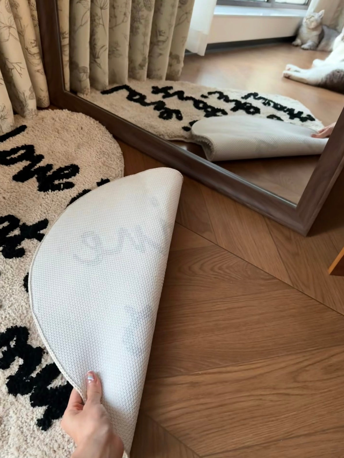 A hand lifts the cream heart rug to show its pale textured backing, with black script visible on the front.