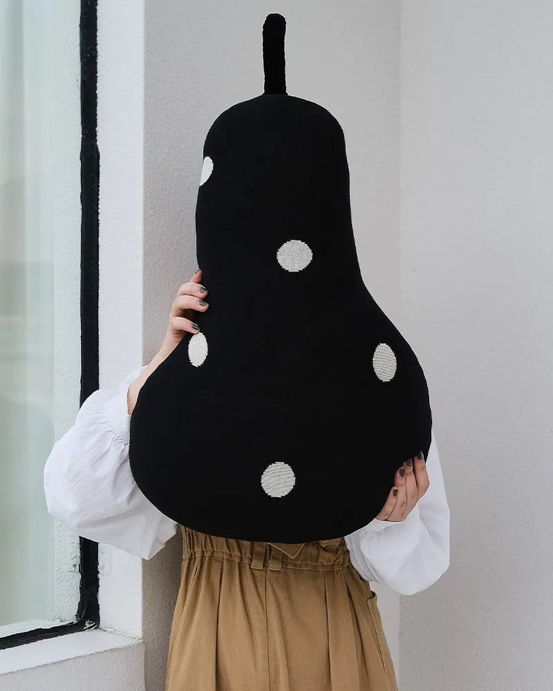 A person wearing a white long-sleeve top and tan skirt holds a large black pear-shaped plush pillow with white polka dots against their face in an indoor setting.