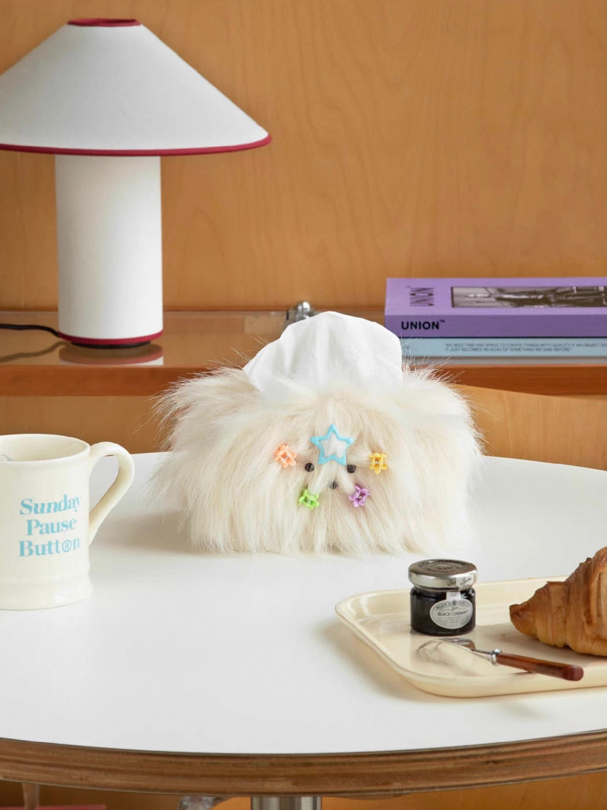 Cream White tissue holder with colorful clips stands on a white tabletop near a lamp and stacked books.
