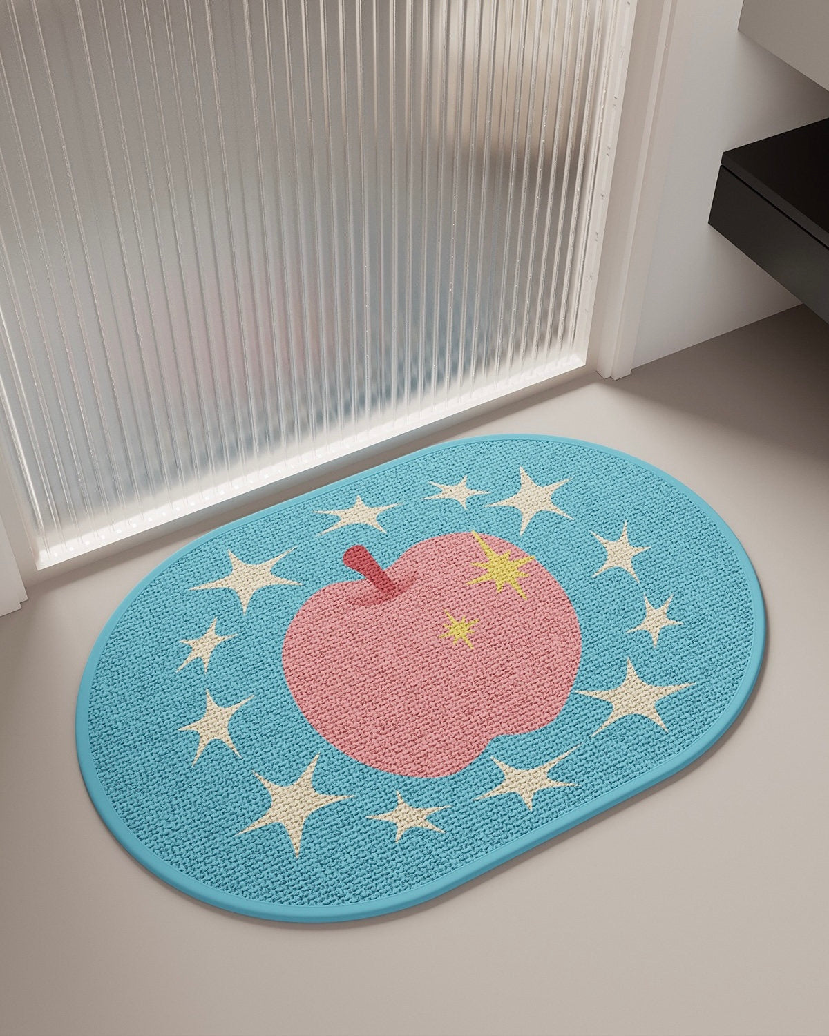 An oval woven doormat with a light blue border, featuring a large pink apple with yellow stars and white stars on a gray floor in front of a ribbed frosted glass door.