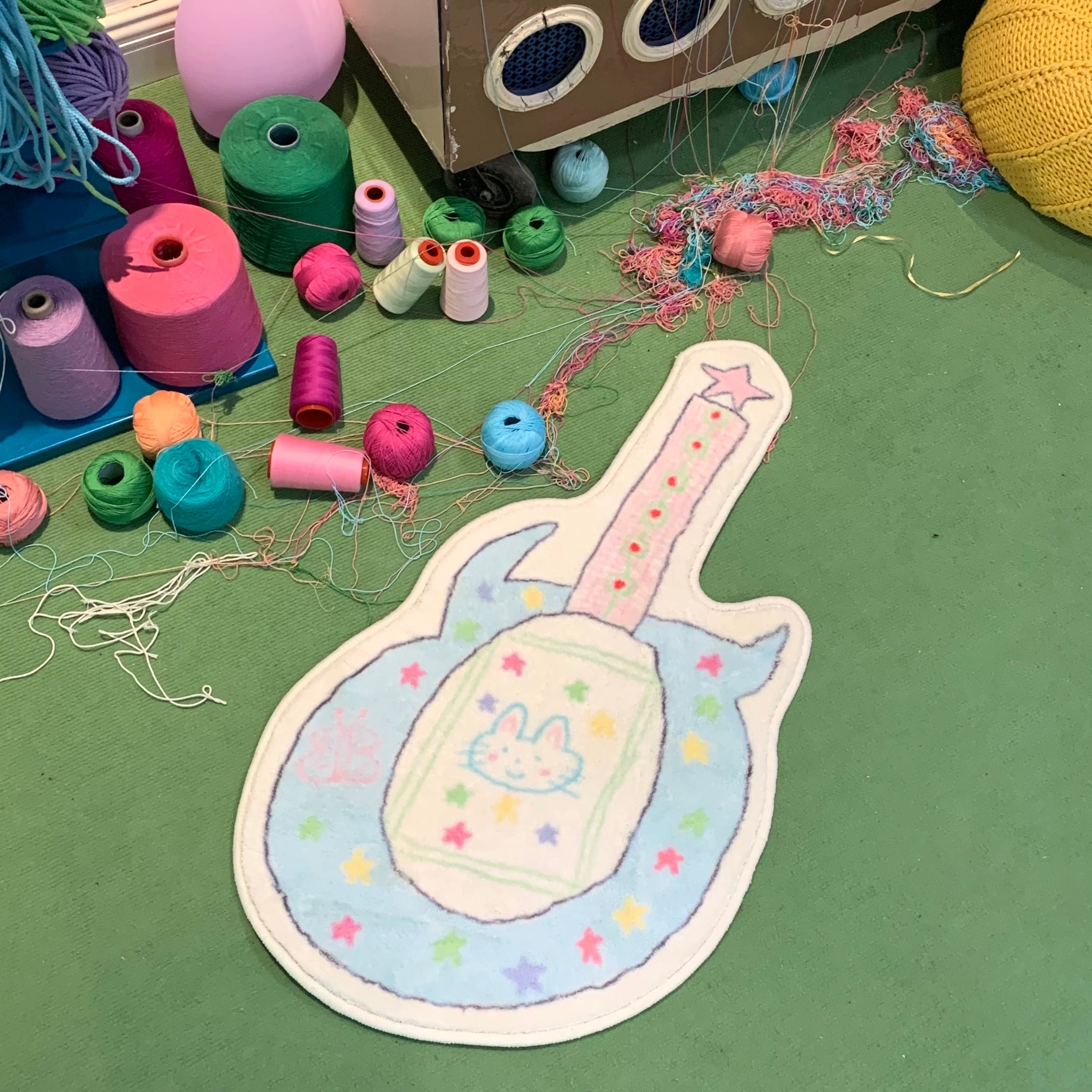 A guitar-shaped plush rug with a bunny design and colorful embroidered stars, displayed on a green floor surrounded by colorful yarn skeins and a cardboard box.