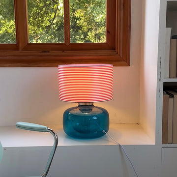 Table lamp with blue base and pink striped shade on a white shelf next to a window.