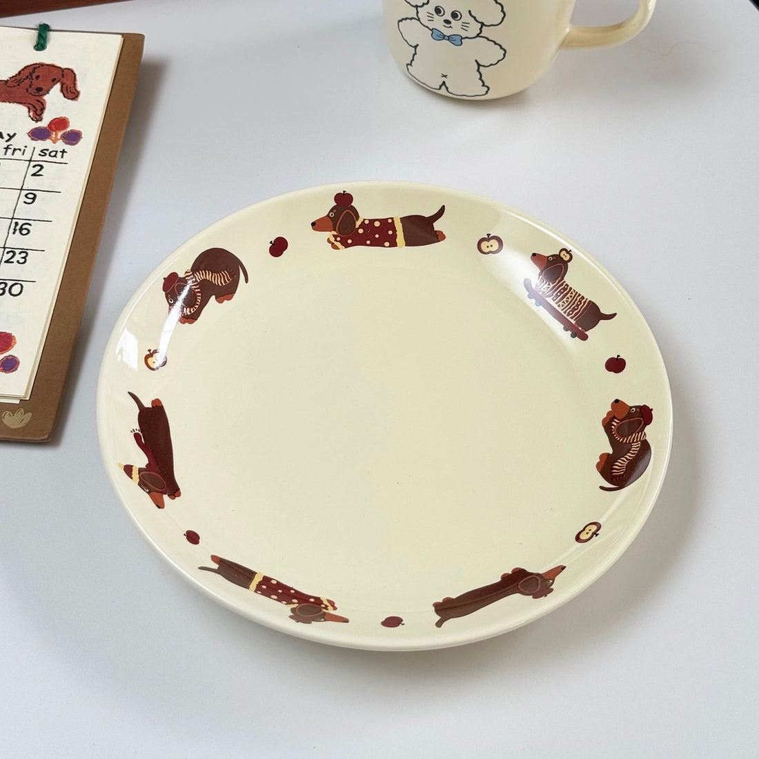 A pretzel cookie on a cream-colored ceramic plate decorated with brown dachshunds, set on a white table next to a mug and a calendar.