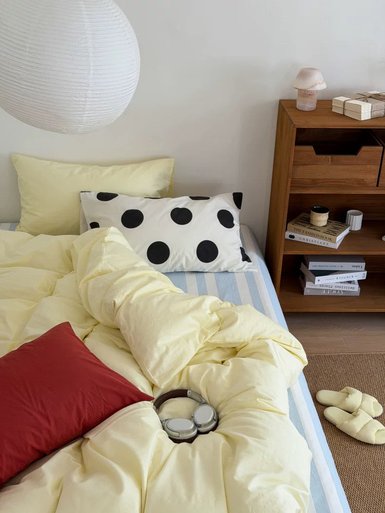 A cozy bedroom scene showing a bed made with pale yellow bedding, a striped sheet, a black-and-white polka dot pillow, a red accent pillow, and a pair of over-ear headphones resting on the duvet. A wooden nightstand, white wall, and floor are visible in the room.
