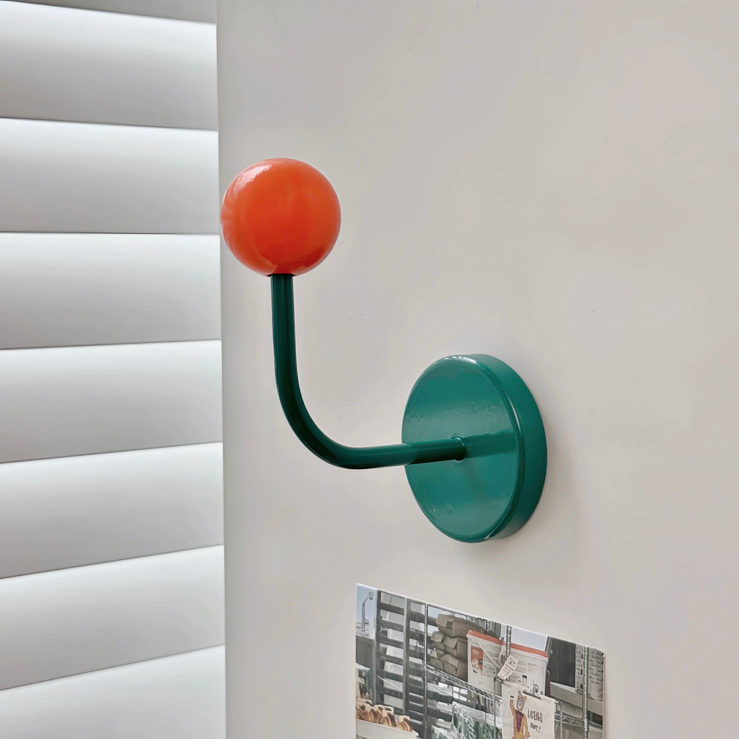 A modern wall-mounted coat rack hook with a round orange top and a curved dark green stem attached to a round green wall plate, shown mounted on a white wall next to window blinds.