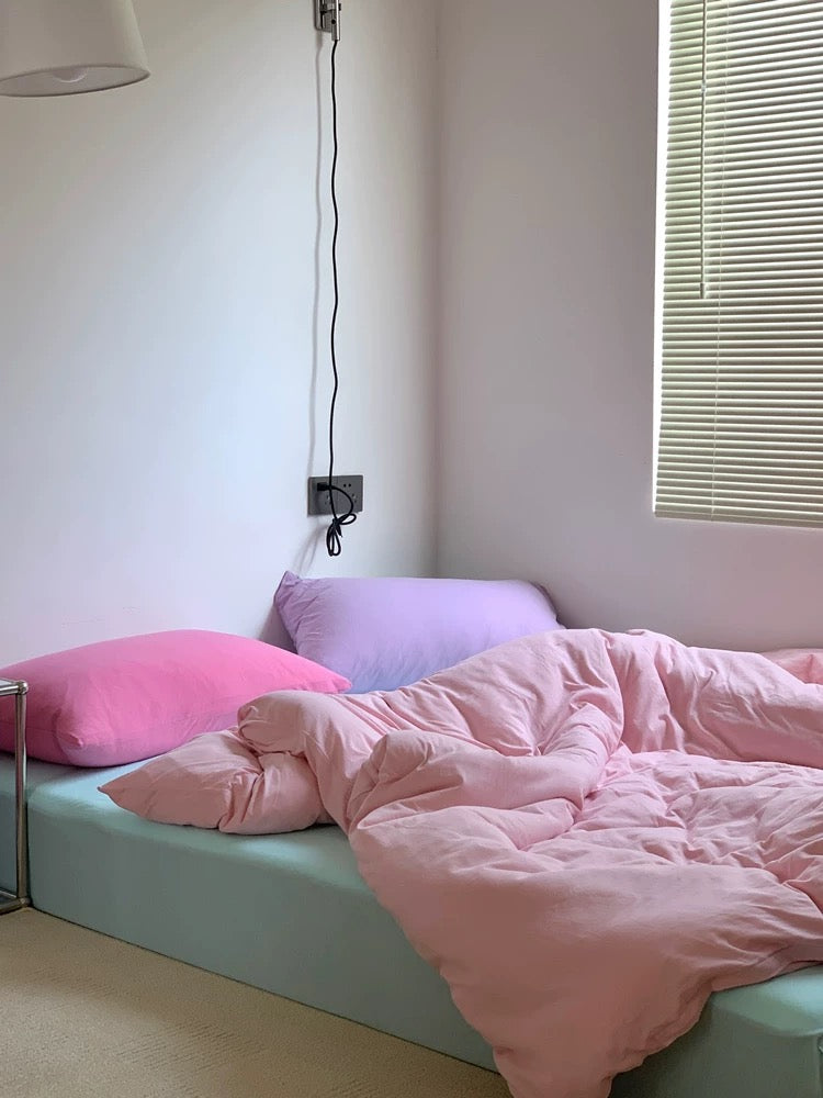 Bed with pink bedding and pillows, a small table with a teapot and bowl on a light blue bedspread.