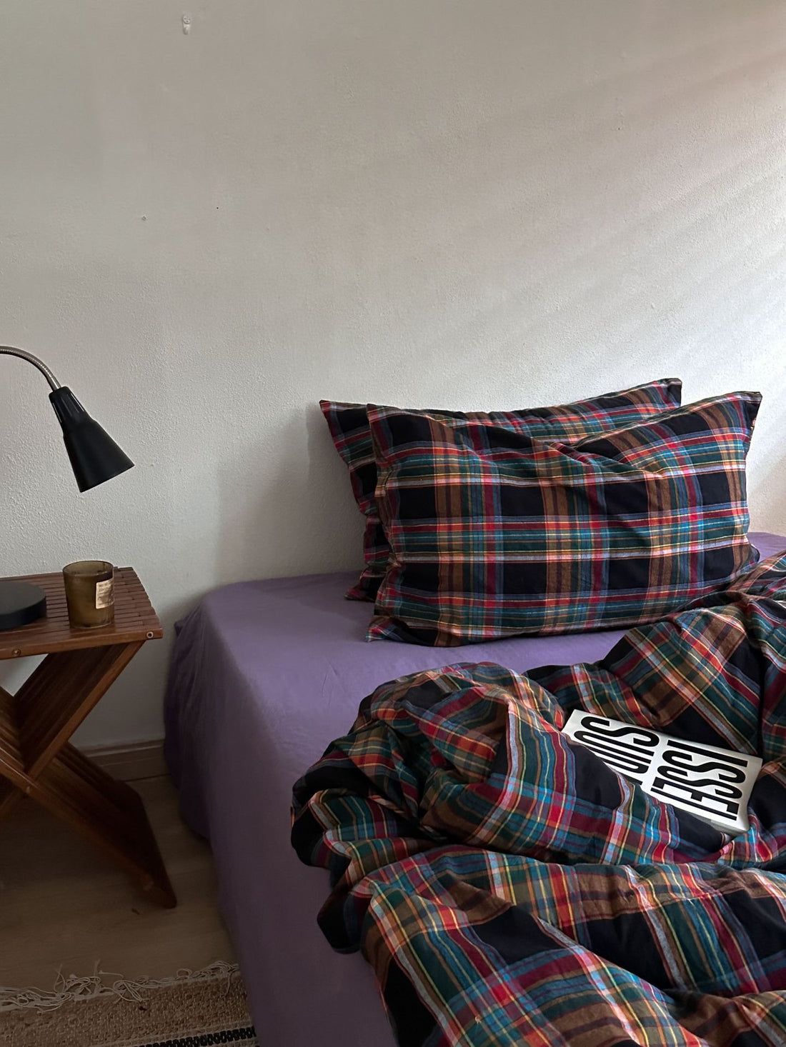 A bed in a bedroom styled with a multicolor plaid bedding set, featuring a purple sheet underneath. The plaid pattern contains shades of red, green, blue, and black.