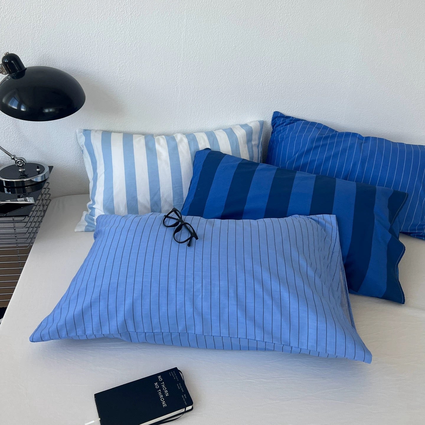 Three blue striped pillowcases arranged on a bed, with a black desk lamp on the left and a black notebook in the foreground.