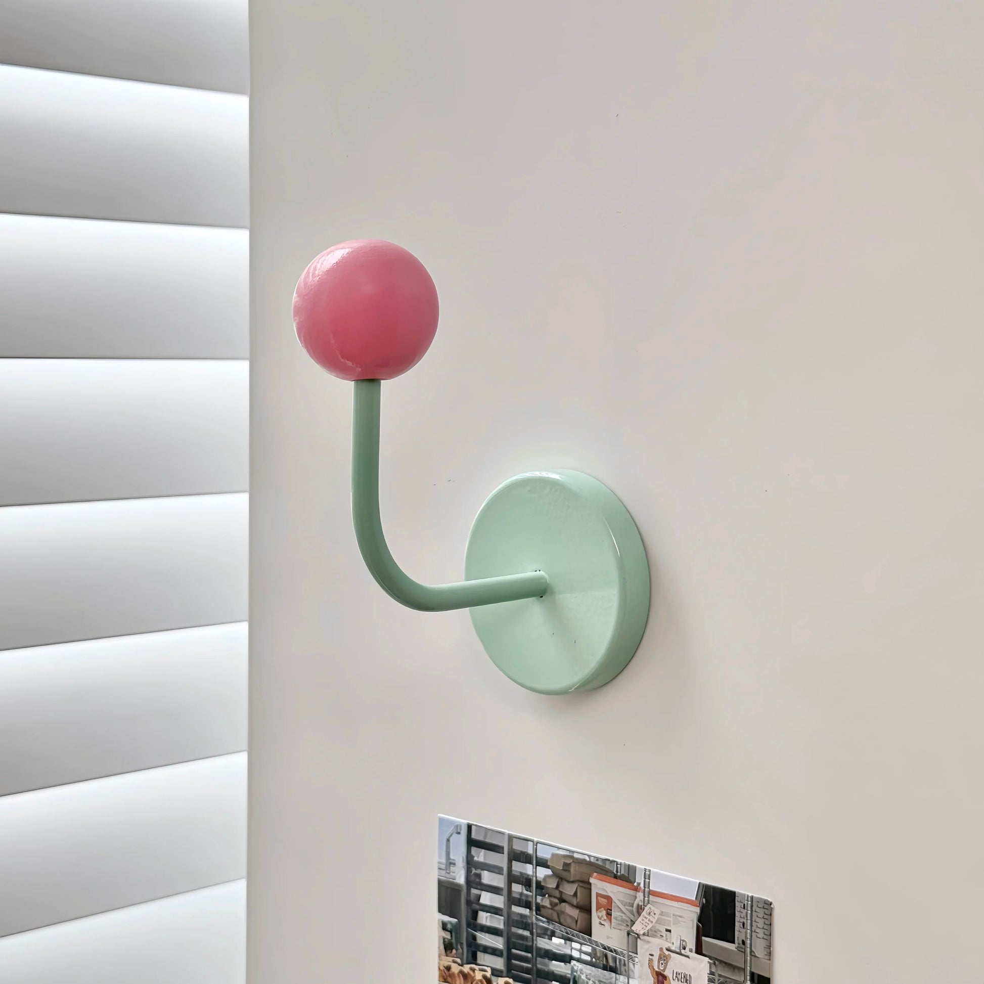 A mint green wall-mounted hook with a pink spherical top, shown attached to a white wall next to window blinds.